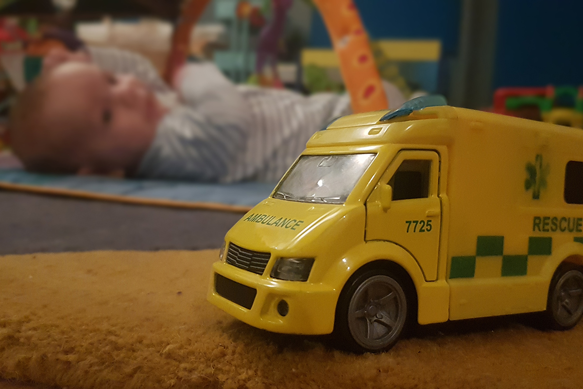 Toy ambulance in focus in foreground with a baby lying on floor in the background.