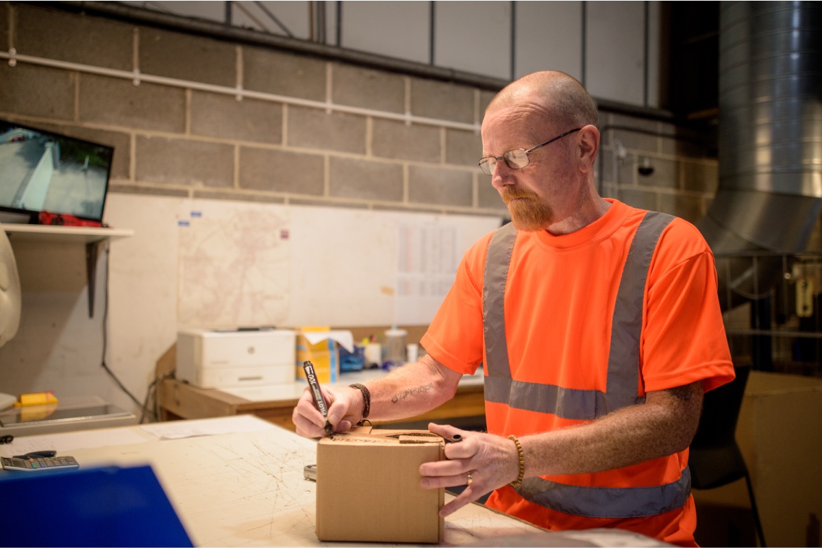 An older male worker in a hi-vis orange jacket seals up a cardboard box