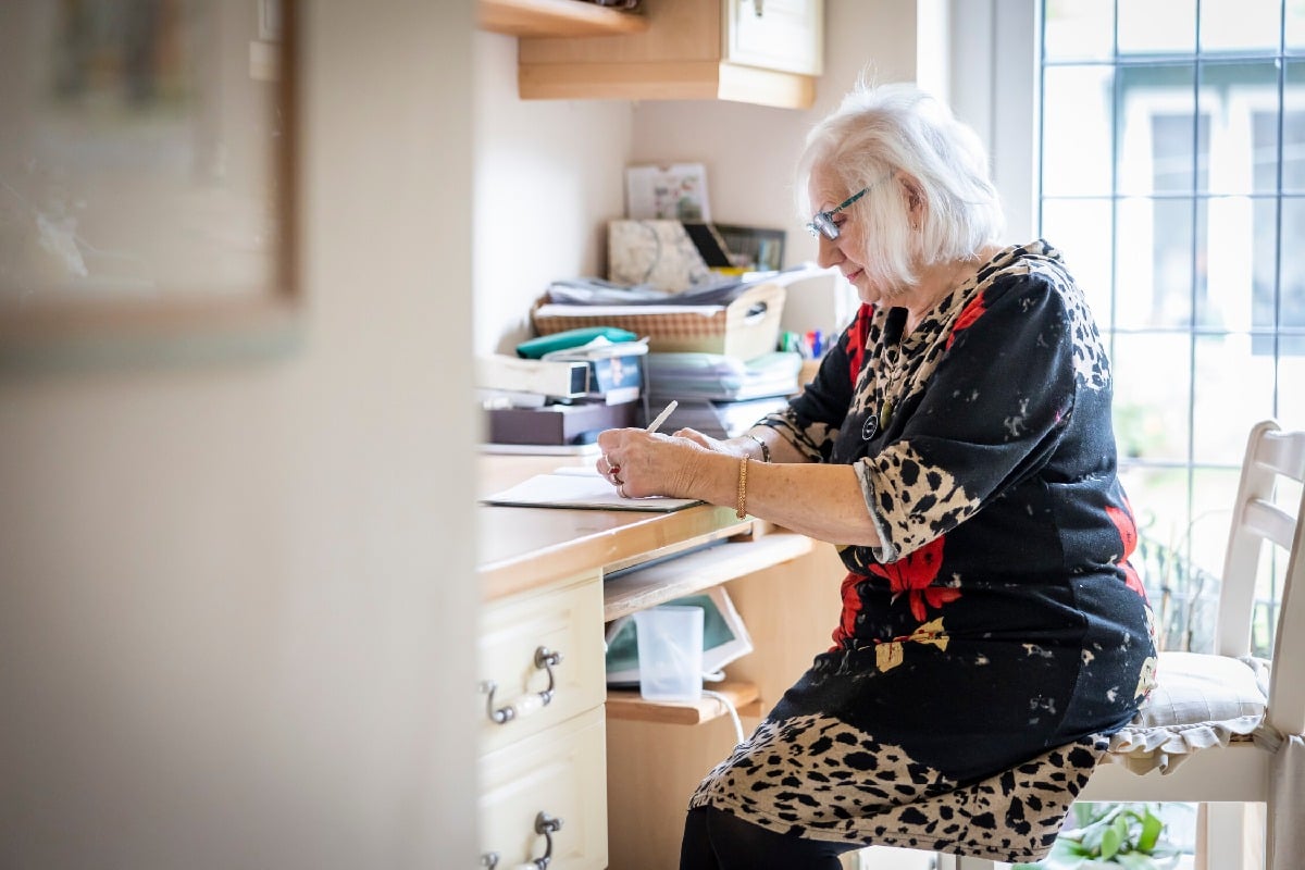 An older woman in a patterned dress writes at a desk in her home