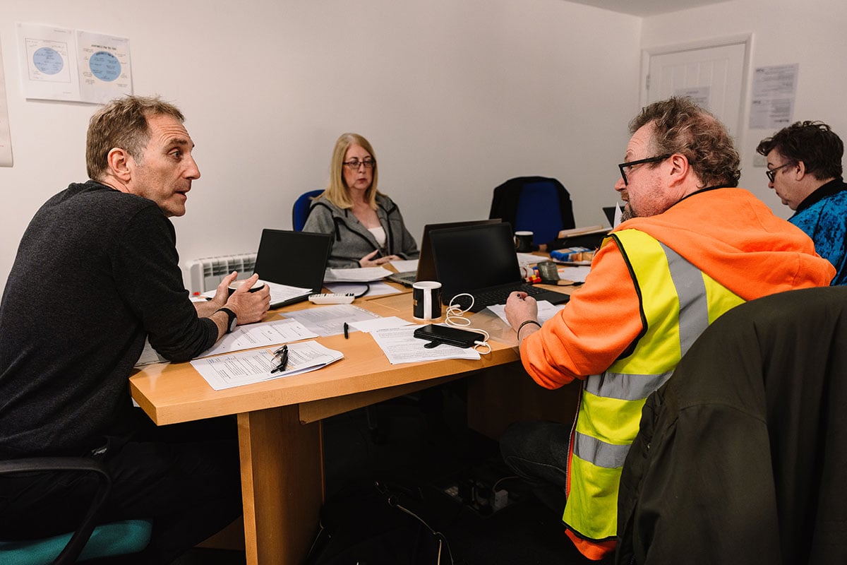 A group of older workers sit around a table and use laptops and write with pen and paper in an employment workshop