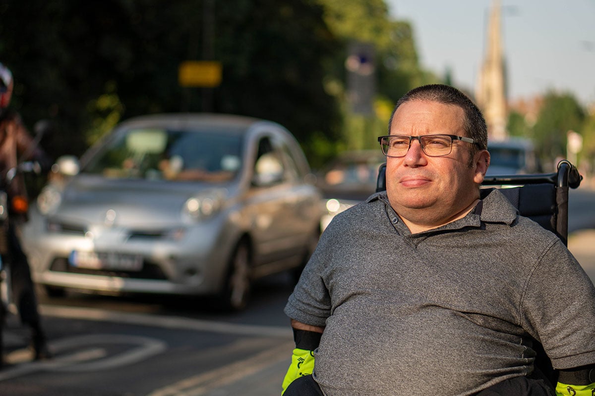 An older worker wearing glasses, a polo shirt, and reflective gloves sits in a wheelchair outdoors, with trees, buildings and cars in the background.