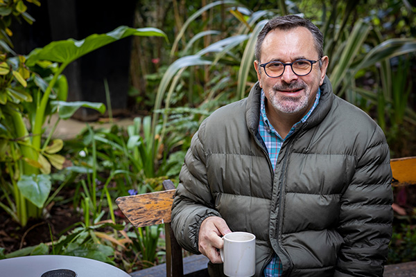 An older man with glasses sits outdoors, holding a cup of tea and looking into the camera.