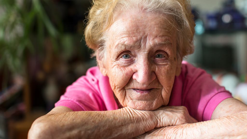 Elderly woman in pink t-shirt resting her chin on crossed arms.