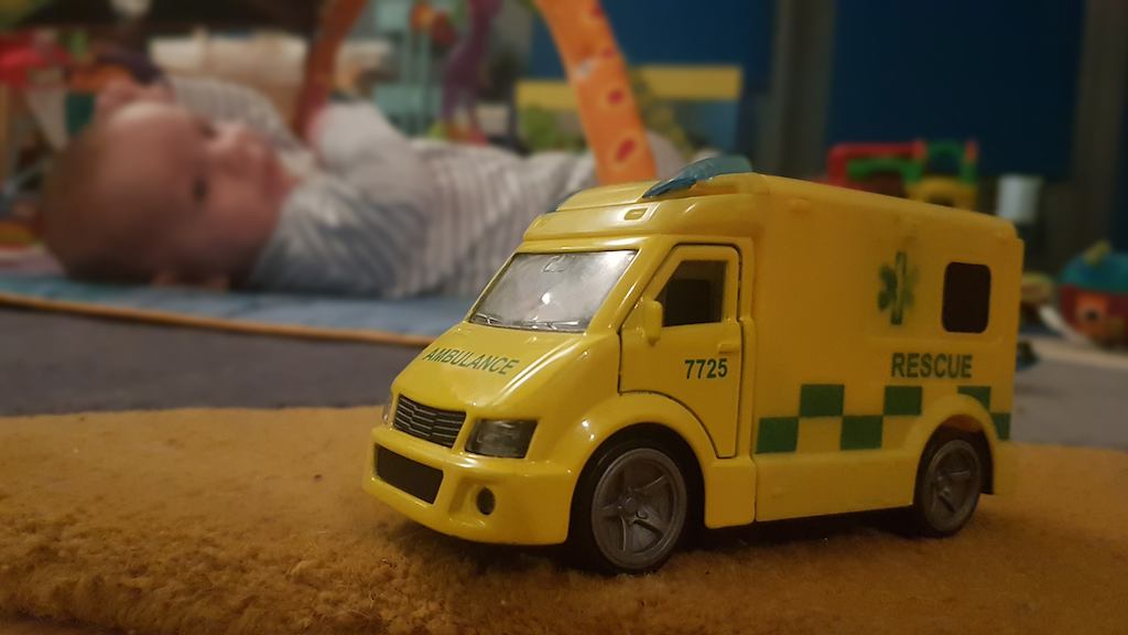 Toy ambulance in focus in foreground with a baby lying on floor in the background.