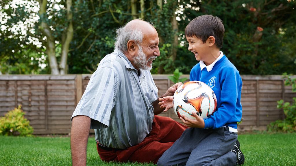Elderly man and grandson sitting on grass with football. 