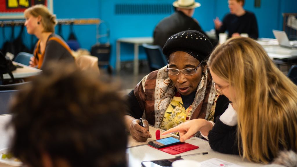Image of a digital skills workshop: an older woman is being taught to use her smartphone by a digital skills volunteer