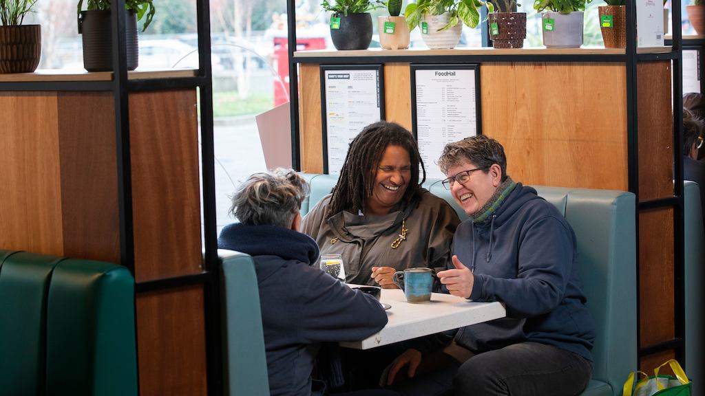 Three older workers having a conversation in a café. The person in the middle is explaining something with their hands, and people on either side are listening. 