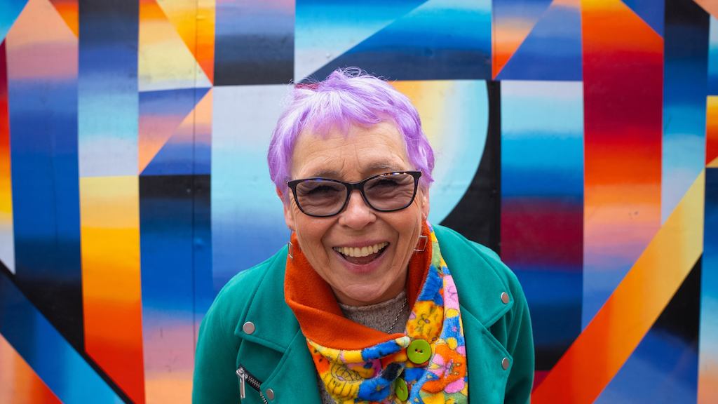 Older woman with purple hair smiling against a colourful background