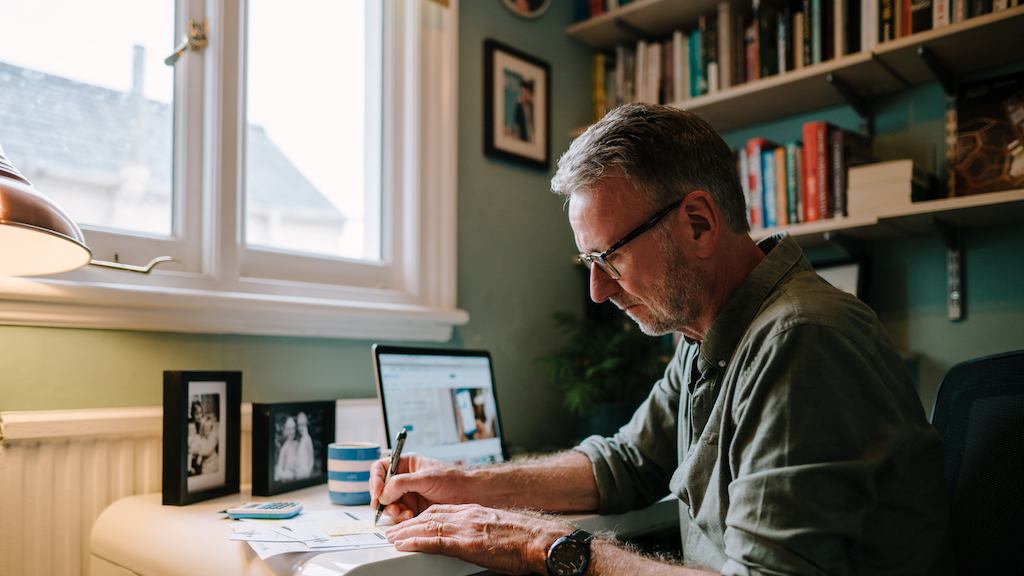 Older man writing in a notebook near his laptop at his desk