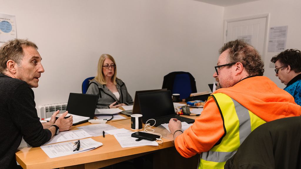 A group of older workers sit around a table and use laptops and write with pen and paper in an employment workshop