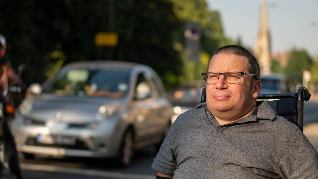 An older worker wearing glasses, a polo shirt, and reflective gloves sits in a wheelchair outdoors, with trees, buildings and cars in the background. 