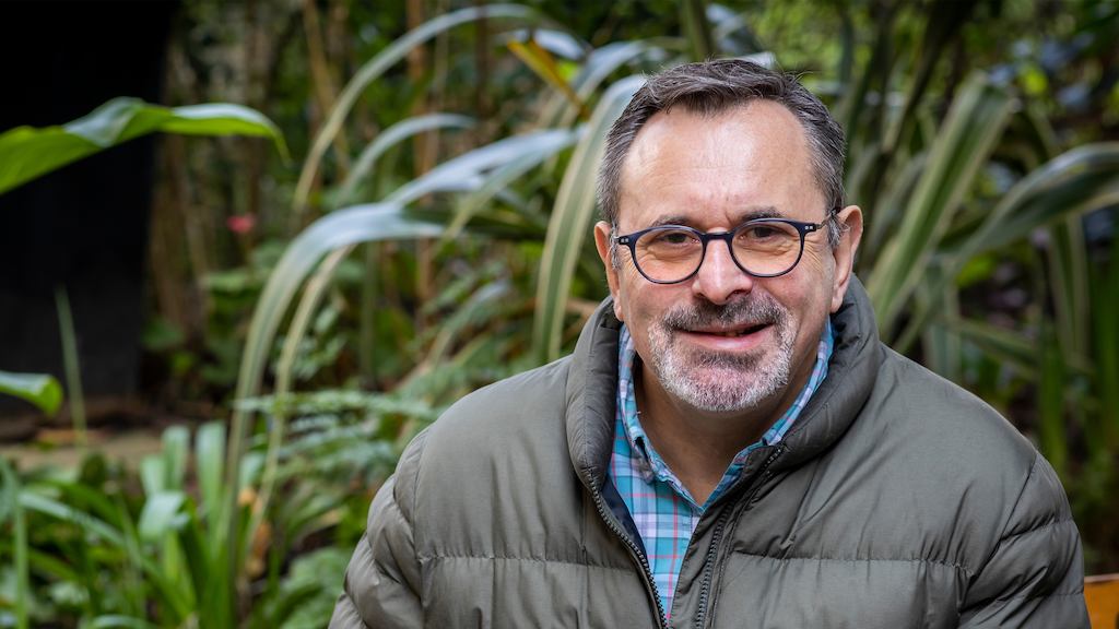 An older man with glasses sits outdoors, holding a cup of tea and looking into the camera. 