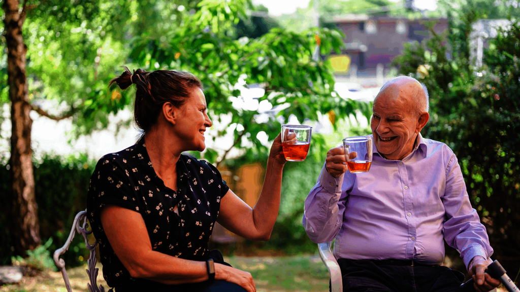 An older man and younger woman sit on chairs in a garden, enjoying cups of tea