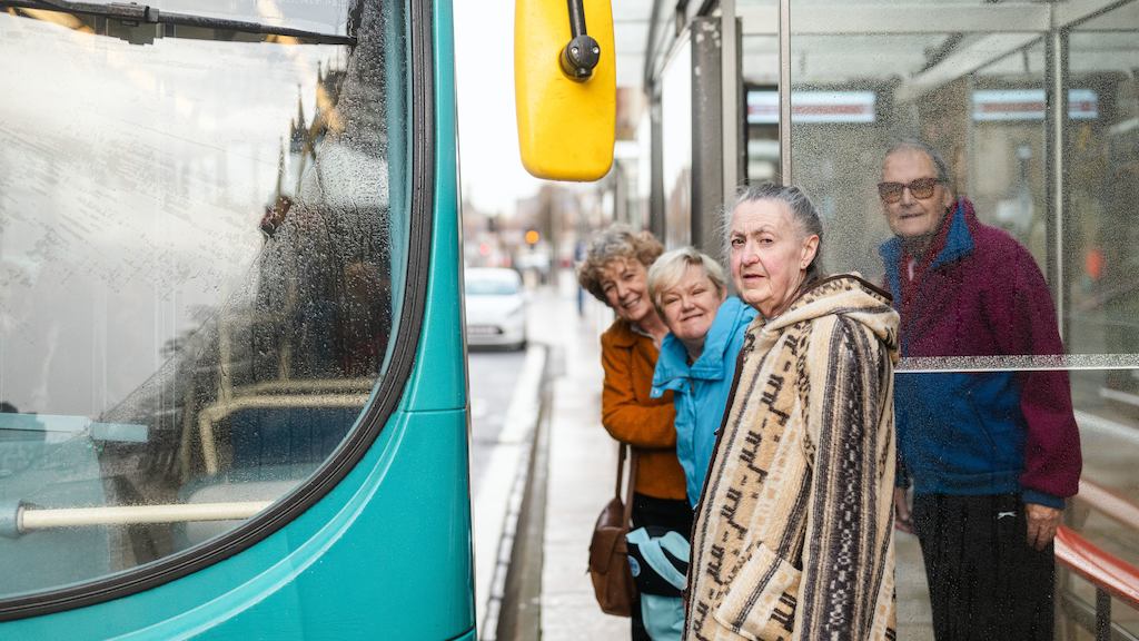A group of older adults stand at a bus stop waiting to board the bus