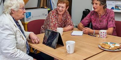 Women gathered at a table having tea and biscuits and using a tablet.