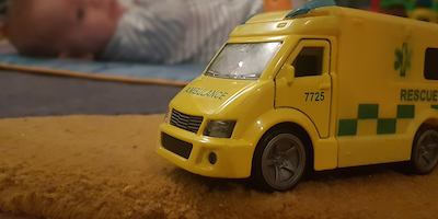 Toy ambulance in focus in foreground with a baby lying on floor in the background.
