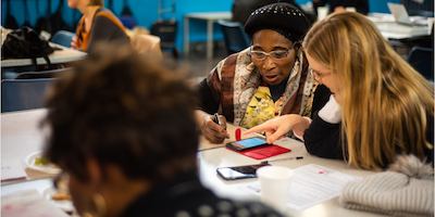 Image of a digital skills workshop: an older woman is being taught to use her smartphone by a digital skills volunteer