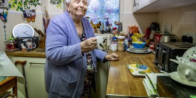 An older woman in a purple cardigan holds a mug of tea in her kitchen