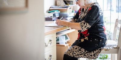 An older woman in a patterned dress writes at a desk in her home