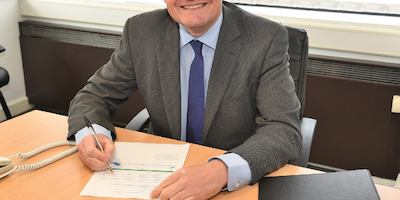 A man wearing a suite smiles whilst writing at a desk