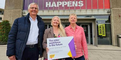 A man and 2 women stood outside a university building, which says 'where change happens' holding a placard which says &quot;The older workforce is the future. that's why University of Essex is working towards becoming an Age-friendly Employer&quot;