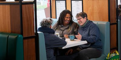 Three older workers having a conversation in a café. The person in the middle is explaining something with their hands, and people on either side are listening.