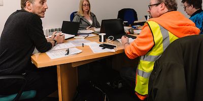 A group of older workers sit around a table and use laptops and write with pen and paper in an employment workshop