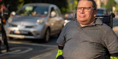 An older worker wearing glasses, a polo shirt, and reflective gloves sits in a wheelchair outdoors, with trees, buildings and cars in the background.