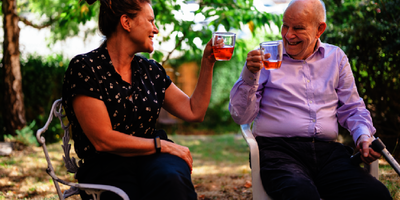 An older man and younger woman sit on chairs in a garden, enjoying cups of tea