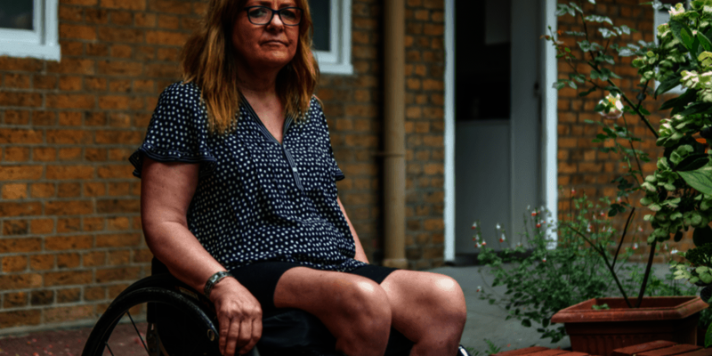 An older woman sits in a wheelchair outside of her house. She is wearing a spotted top and glasses.
