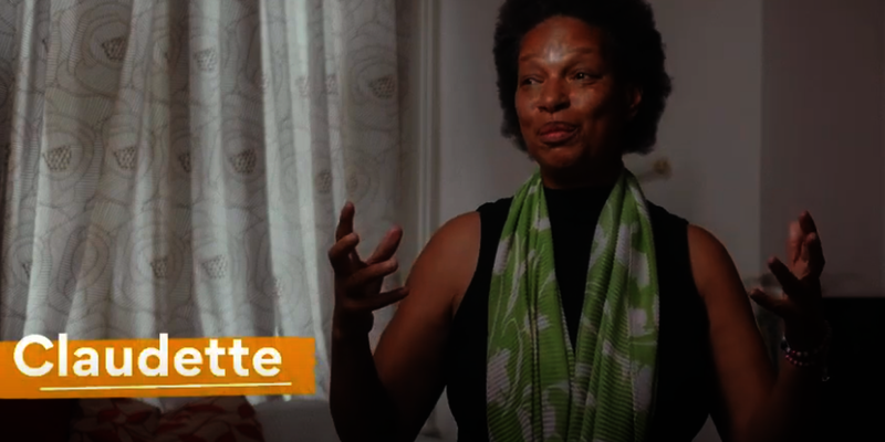 An older person sits in a room. She is wearing a green scarf.