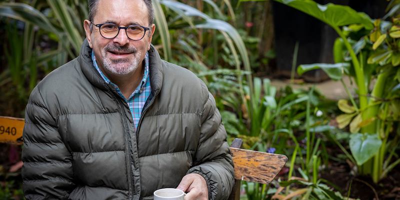 An older man with glasses sits outdoors, holding a cup of tea and looking into the camera. 