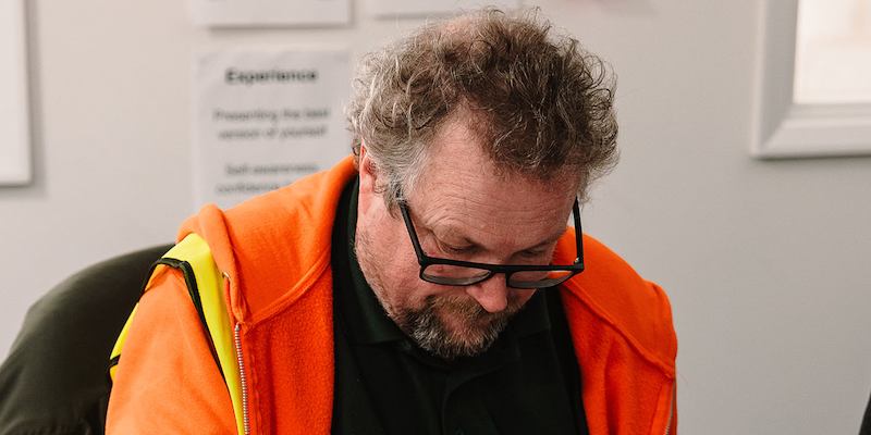 Older worker wearing glasses and a high-visibility vest fills out paperwork at a desk. 