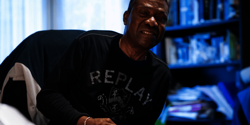 Man working from home, smiling at the camera and wearing a black long-sleeve T-shirt. A full bookshelf is behind him, and paperwork is on the table in front. 