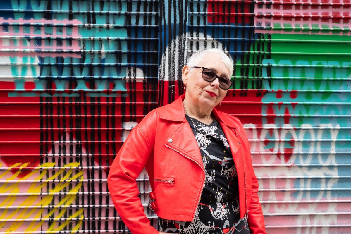 older woman in a red leather jacket poses in front of a colourful wall
