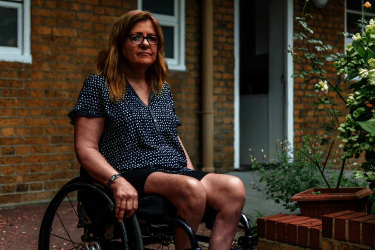 An older woman sits in a wheelchair outside of her house. She is wearing a spotted top and glasses.
