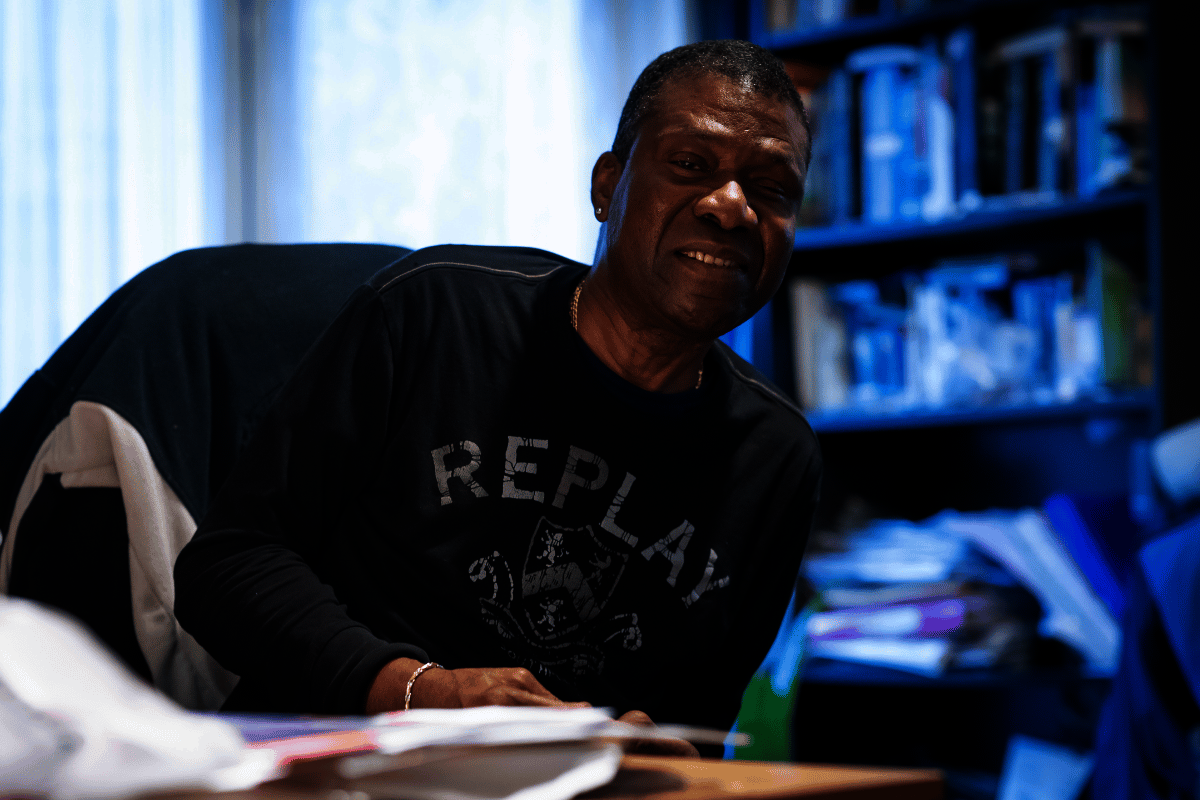 Man working from home, smiling at the camera and wearing a black long-sleeve T-shirt. A full bookshelf is behind him, and paperwork is on the table in front. 
