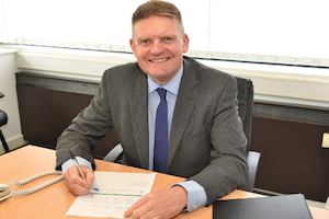 A man wearing a suite smiles whilst writing at a desk