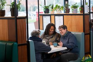 Three older workers having a conversation in a café. The person in the middle is explaining something with their hands, and people on either side are listening.