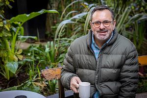 An older man with glasses sits outdoors, holding a cup of tea and looking into the camera.