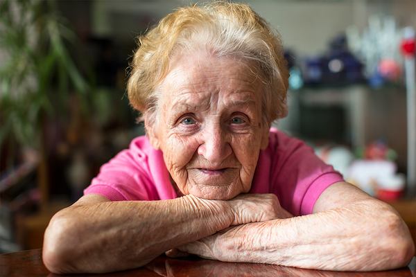 Elderly woman in pink t-shirt resting her chin on crossed arms.