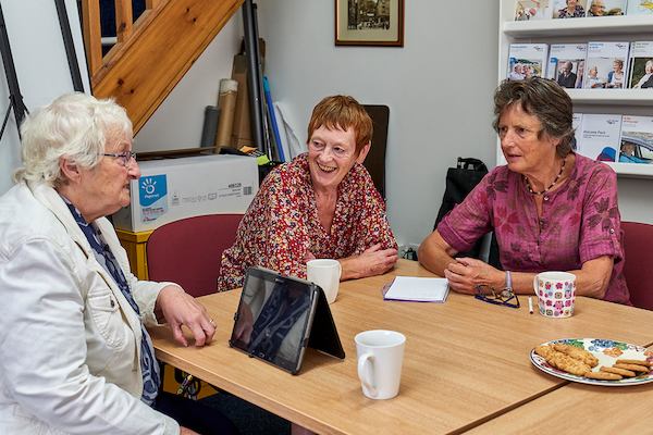 Women gathered at a table having tea and biscuits and using a tablet.