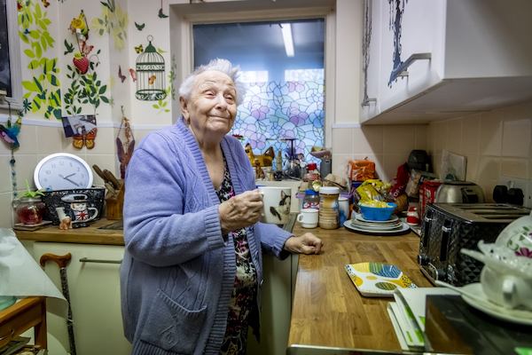 An older woman in a purple cardigan holds a mug of tea in her kitchen