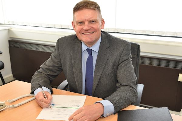 A man wearing a suite smiles whilst writing at a desk