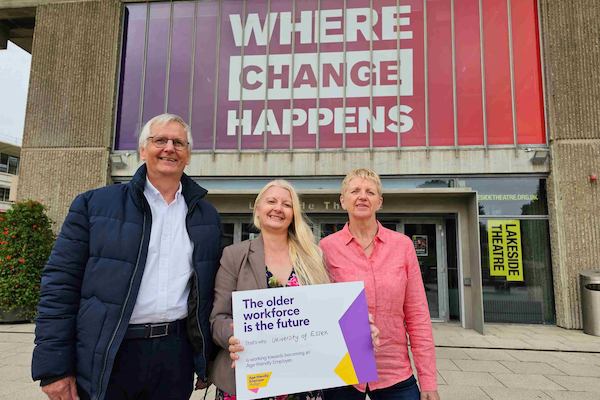 A man and 2 women stood outside a university building, which says 'where change happens' holding a placard which says "The older workforce is the future. that's why University of Essex is working towards becoming an Age-friendly Employer"