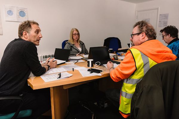 A group of older workers sit around a table and use laptops and write with pen and paper in an employment workshop