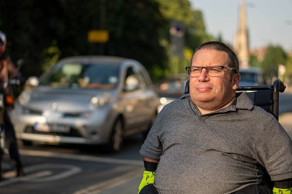 An older worker wearing glasses, a polo shirt, and reflective gloves sits in a wheelchair outdoors, with trees, buildings and cars in the background.