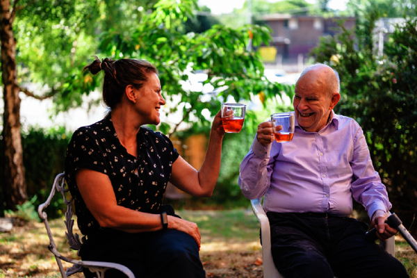 An older man and younger woman sit on chairs in a garden, enjoying cups of tea
